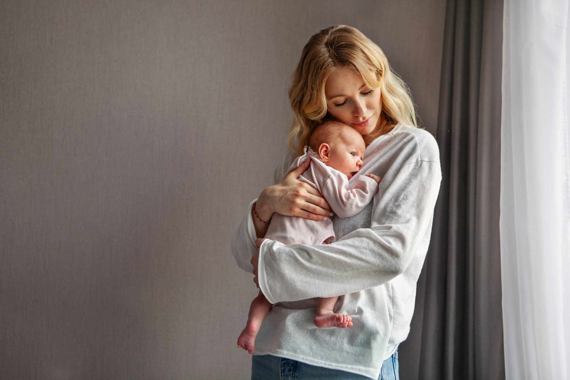 Young beautiful mother holds newborn baby while standing at home near window