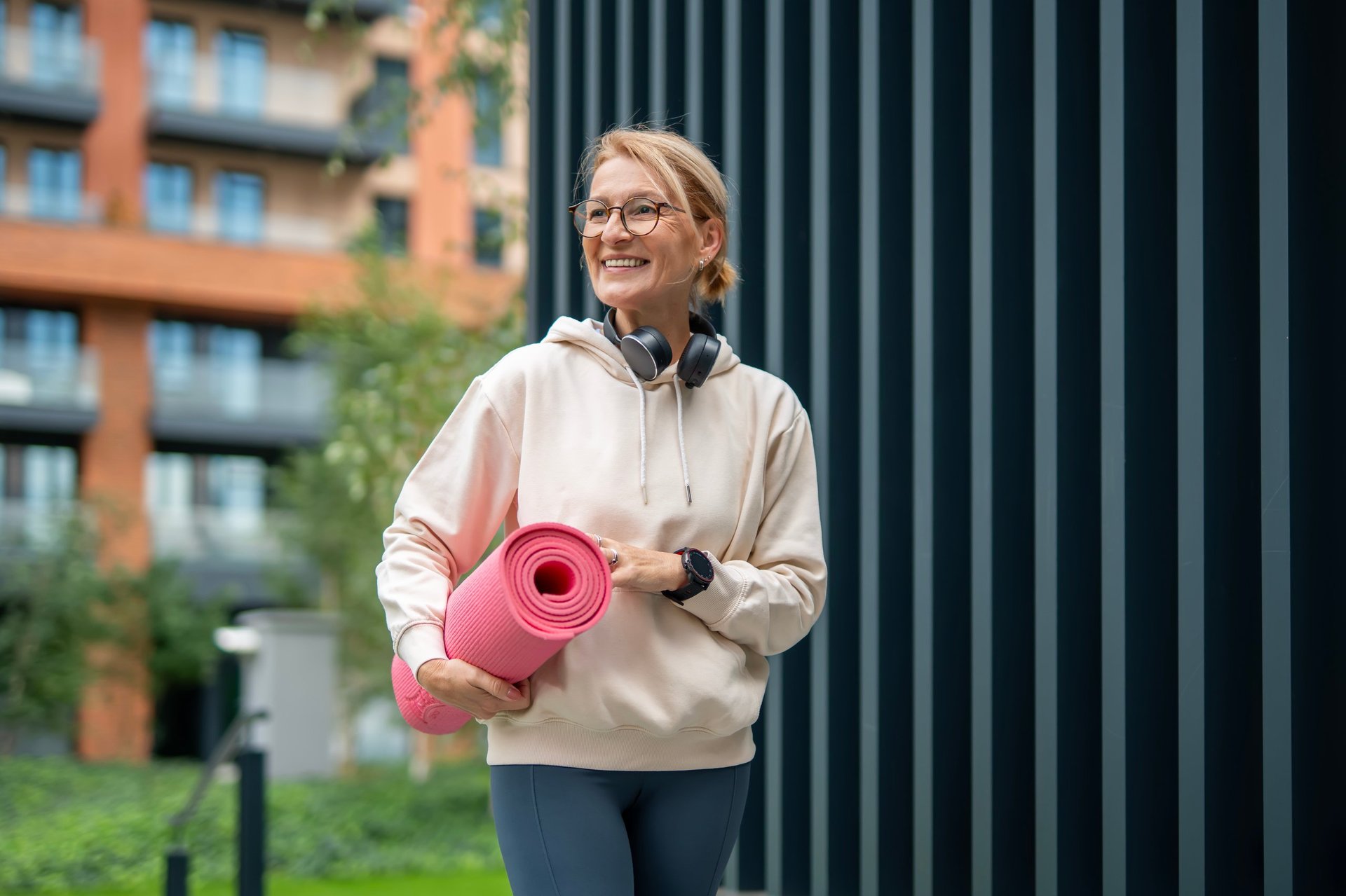 Mature woman smiling and walking through the city with headphones around her neck and a yoga mat over her shoulder